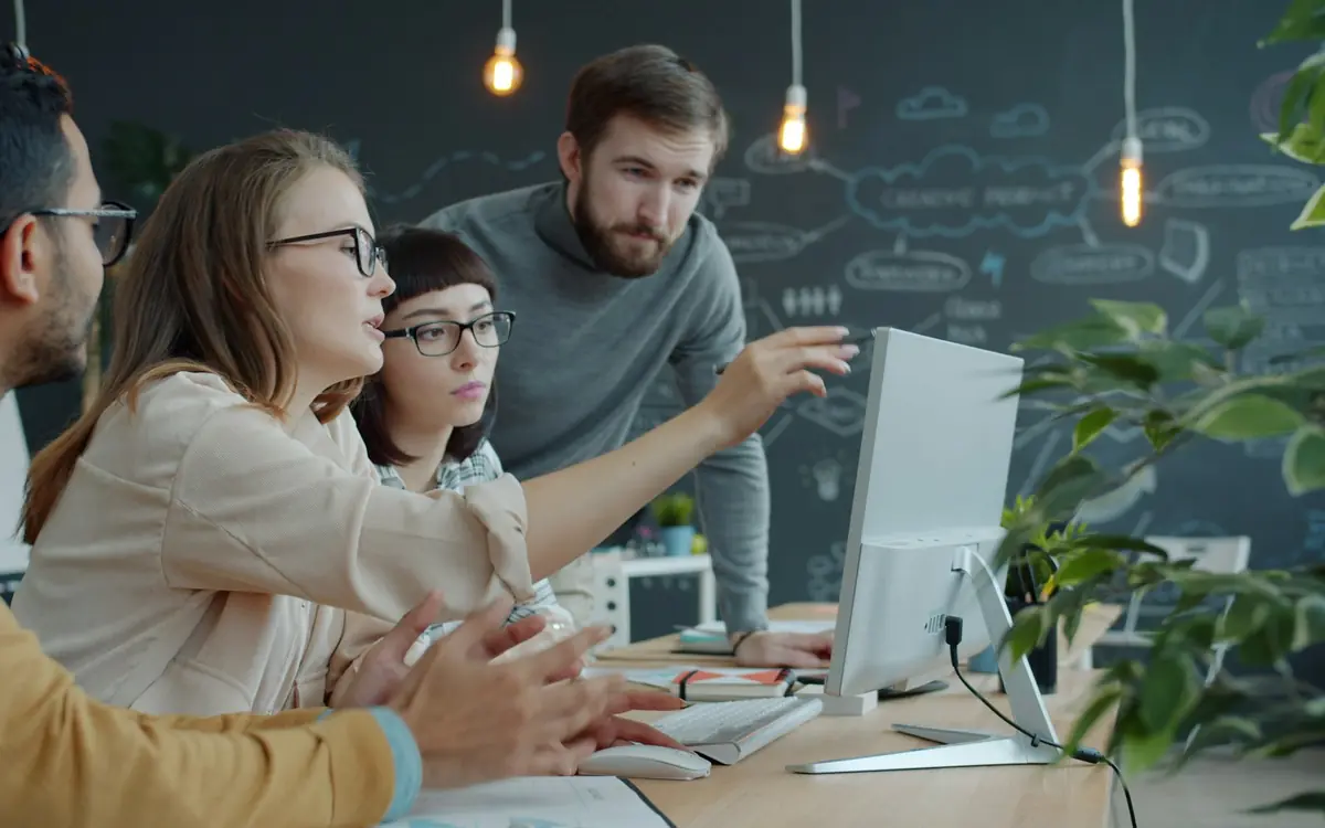 Three colleagues in an open-plan office pointing at one shared computer screen while reviewing business data together