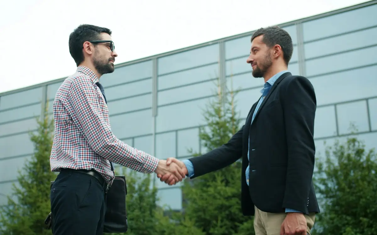Two businessmen in smart clothes shaking hands outdoors in front of a modern building after agreeing a deal
