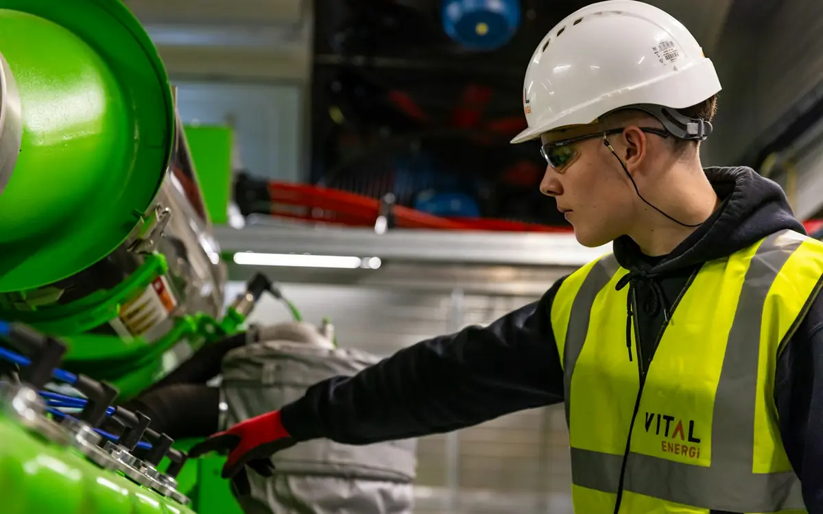 Machine operator in a white hard hat, safety glasses and high-visibility vest checking industrial equipment on a factory floor