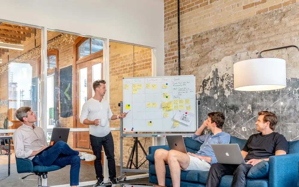 Four men in a brick-walled office, one presenting a plan at a whiteboard covered with sticky notes while three colleagues follow on laptops