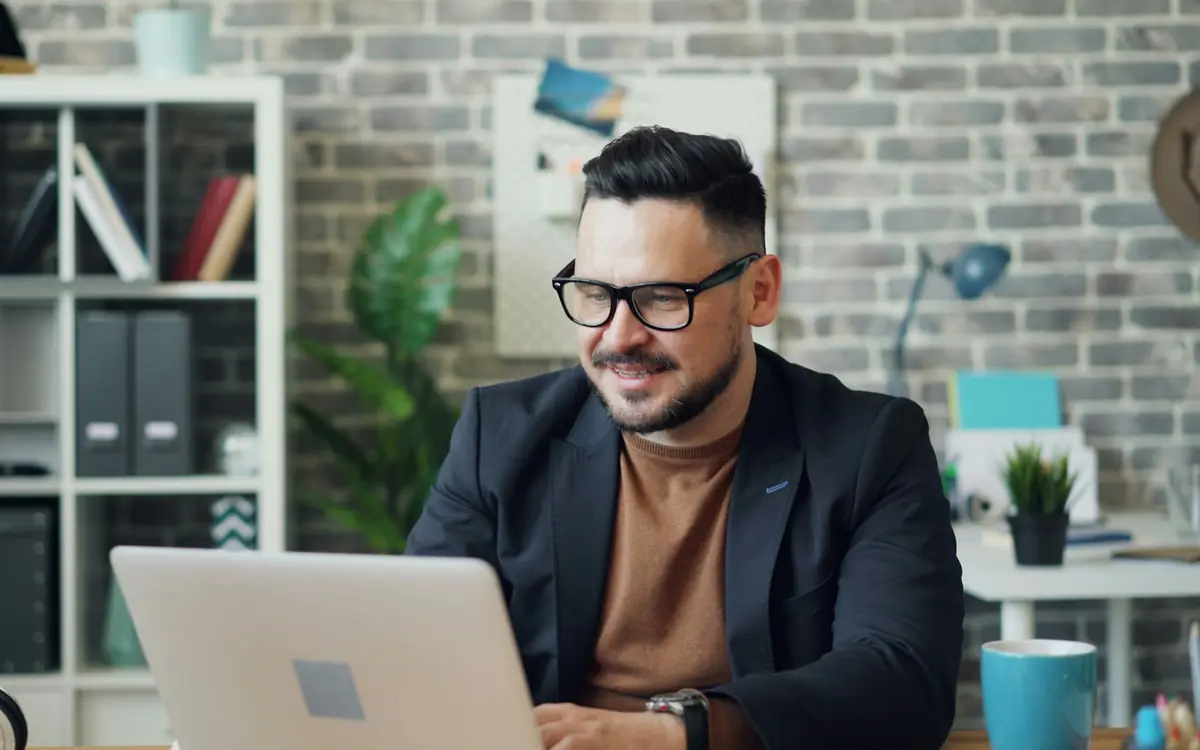 HR manager in a jacket and glasses smiling while reviewing employee records on a laptop at his office desk